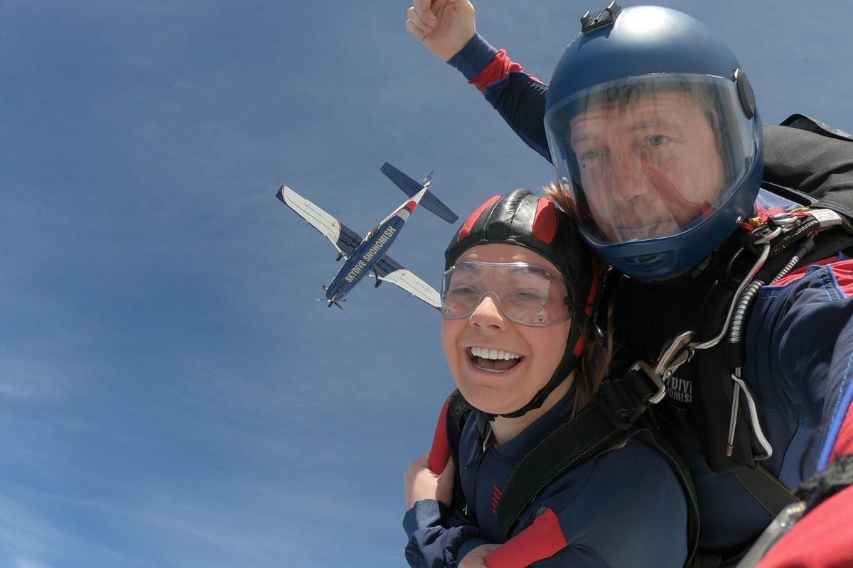 Female tandem student with brown hair smiling while skydiving with Blue, White, and Red airplane above her.